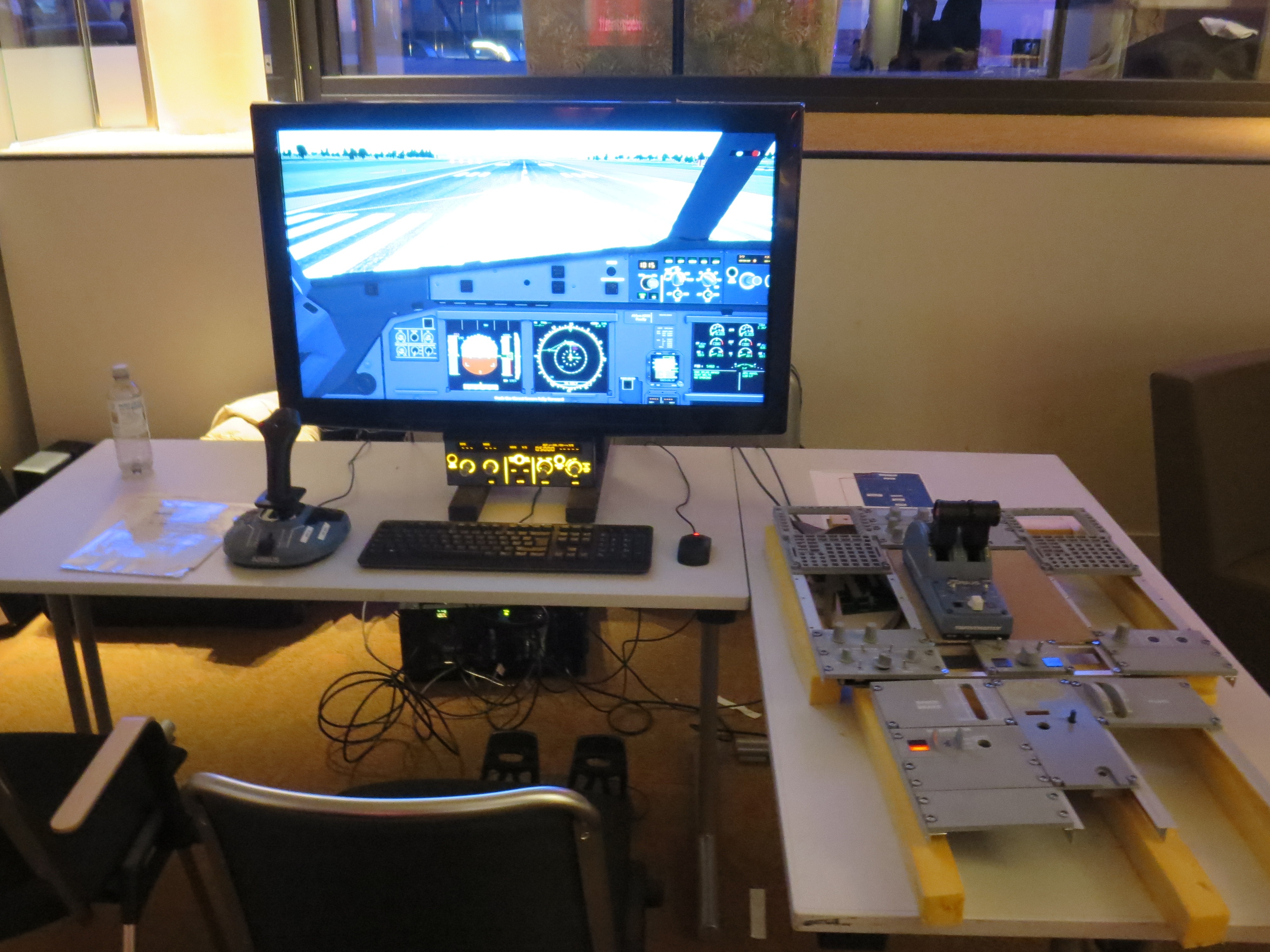 The setup at last years event. The parts of the pedestal are on the right, on a table, on a desk is a bigger screen and in front of it a side stick, keyboard and the control panel for autopilot. On the floor are rudder pedals. In the back are a lot of cables hanging down which lead to the PC